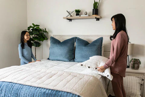 
      A mother and daughter making the bed together using neutral zipper bedding and blue throw pillows and blankets.
    