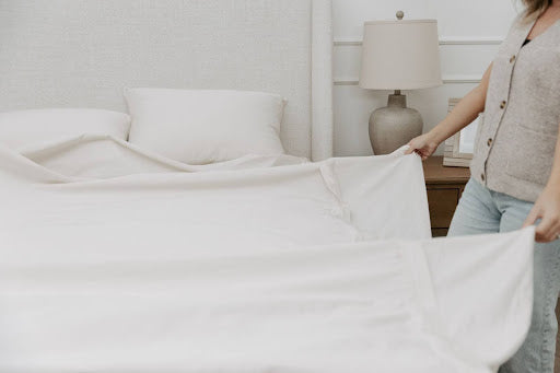 
      A woman arranging white zipper sheets on a bed with a fabric headboard.
    