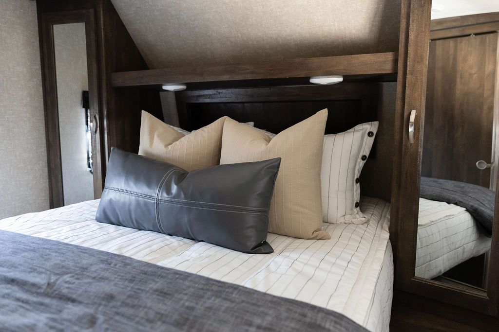 A bed in an RV made with white and pinstripes zipper bedding and neutral and leather pillows and blanket.