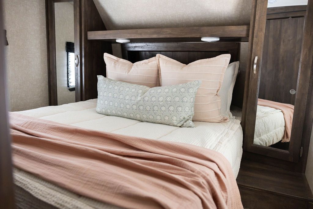 A bed in an RV made with cream waffle textured zipper bedding with an assortment of pink and green pillows and blanket.