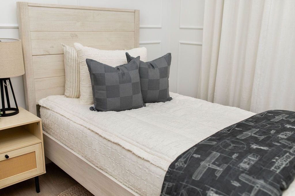 A twin bed made with cream textured zipper bedding and an assortment of neutral throw pillows and an airplane pattern black blanket.
