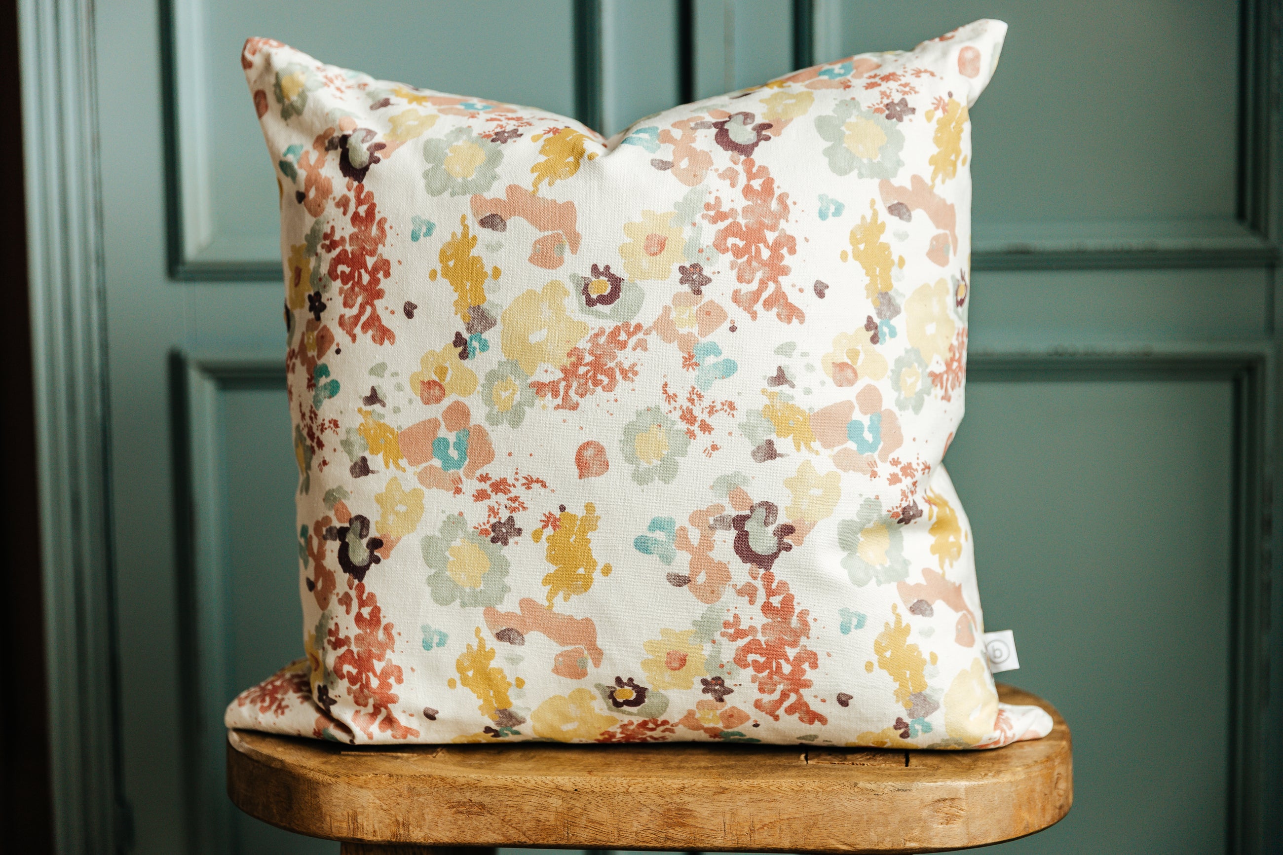 A floral throw pillow on a wooden stool in front of a green wall.