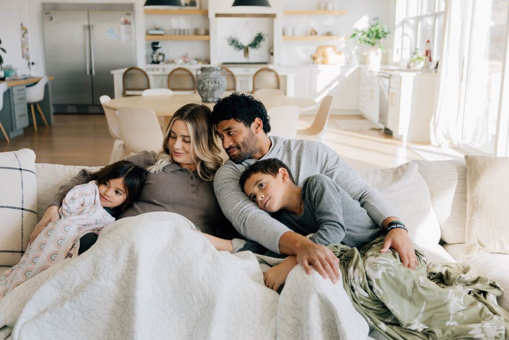 A family consisting of a mom, a dad, a son and a daughter sitting on the couch together, all wrapped up in their blankets and cuddling.