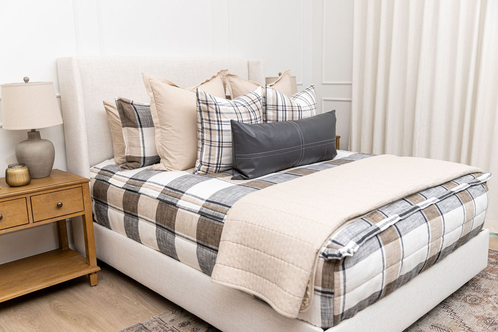 A bed made with tan, cream, and charcoal zipper bedding and an assortment of neutral pillows and blanket.