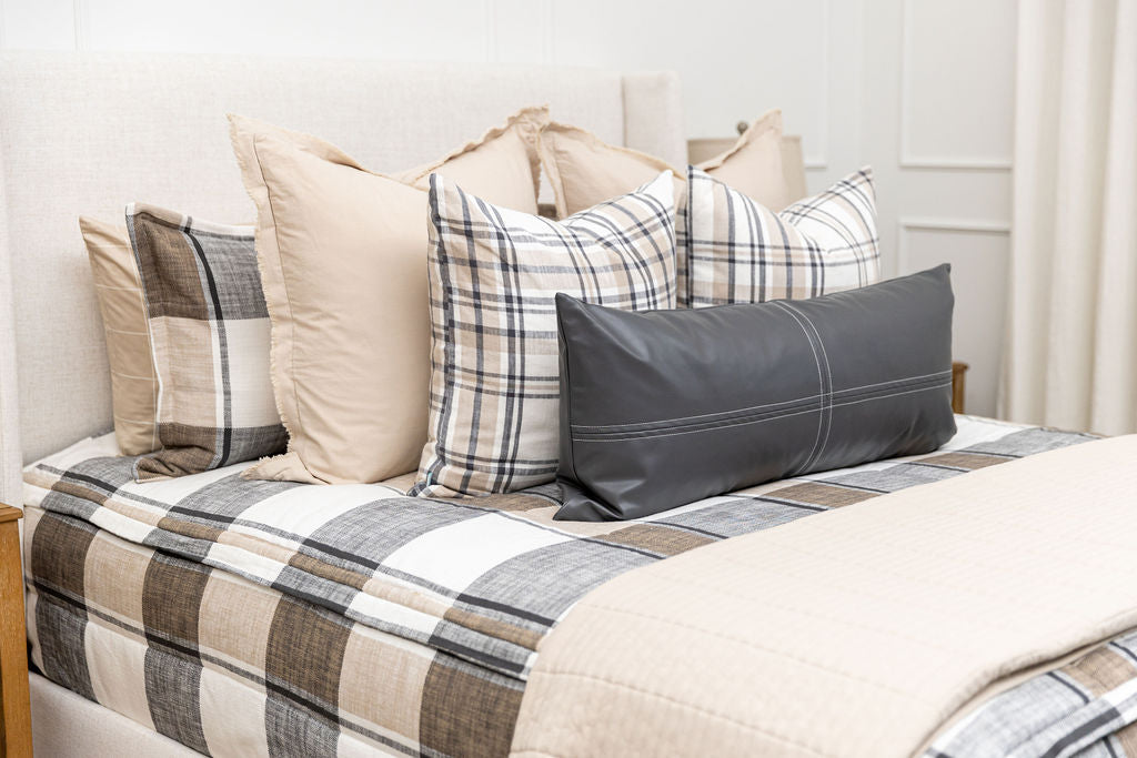 A close up view of a bed made with tan, cream, and charcoal zipper bedding and an assortment of neutral pillows and blanket.
