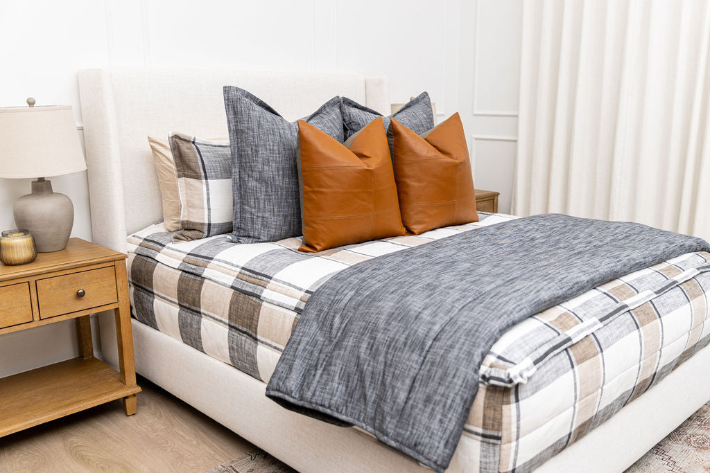 A bed made with charcoal, cream, and tan plaid zipper bedding and an assortment of dark gray and leather throw pillows and matching blanket.