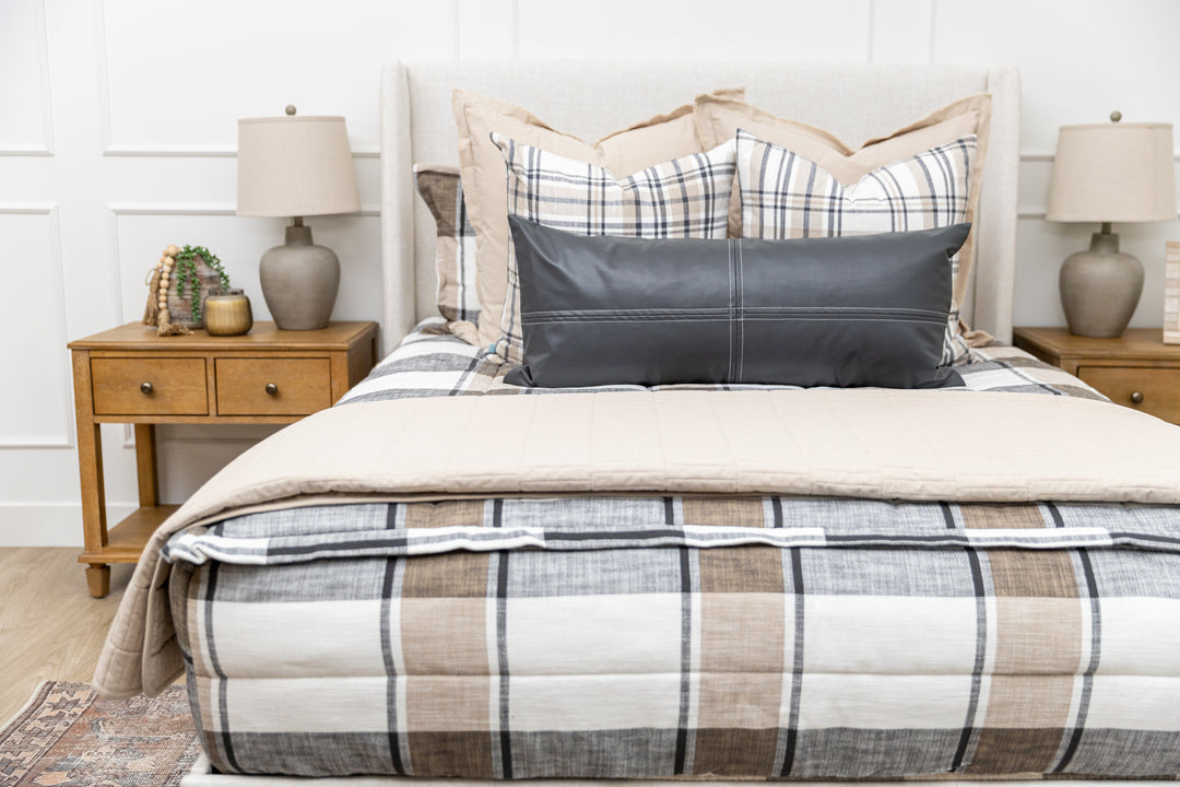 A bed made with cream, charcoal, and tan plaid zipper bedding and an assortment of neutral and leather throw pillows and blankets.