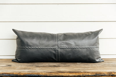 A faux leather charcoal lumbar pillow sitting on a brown bench with a white backdrop.