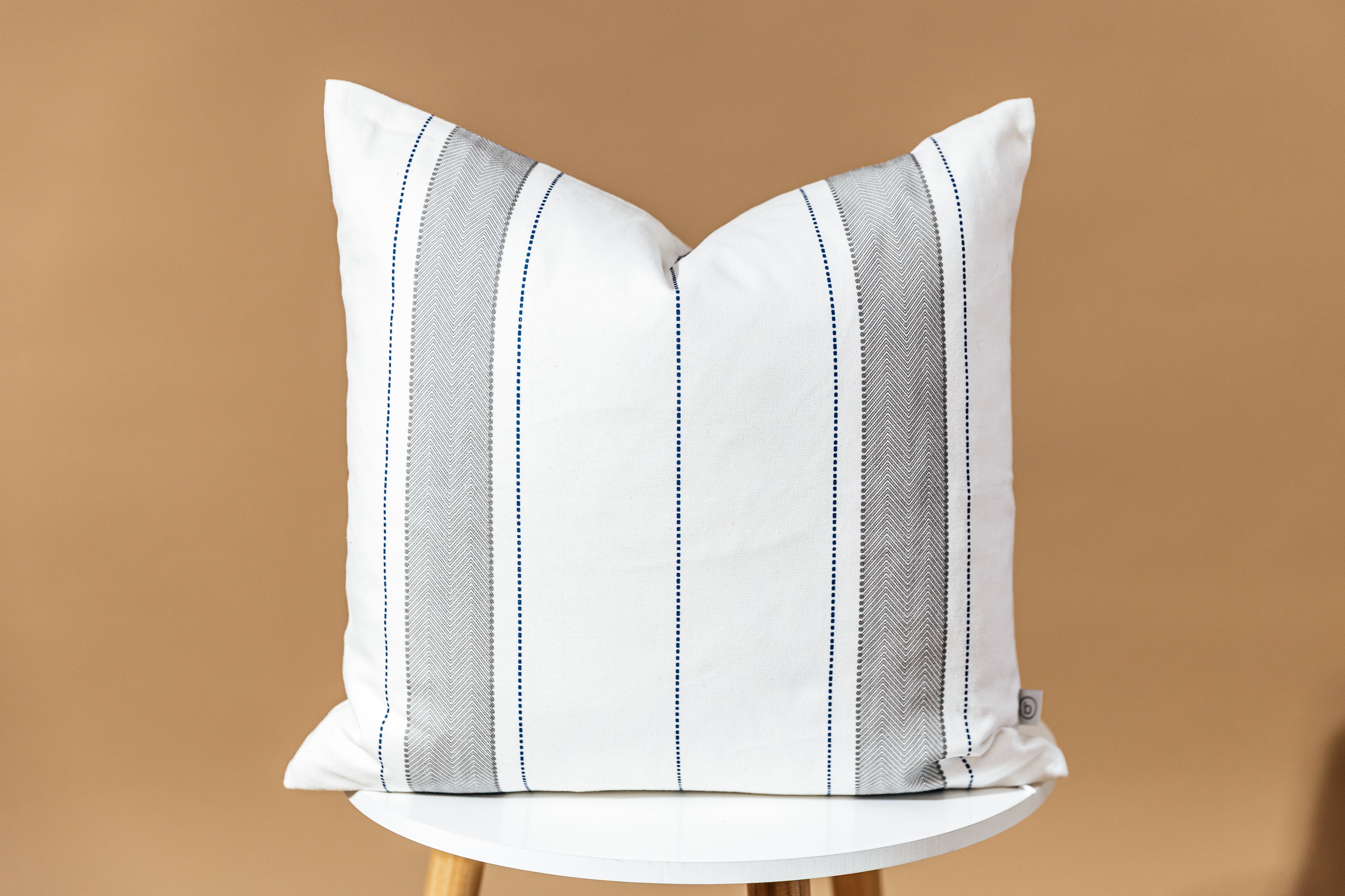 A white, gray, and navy striped pillow sitting on a stool in front of a brown backdrop.