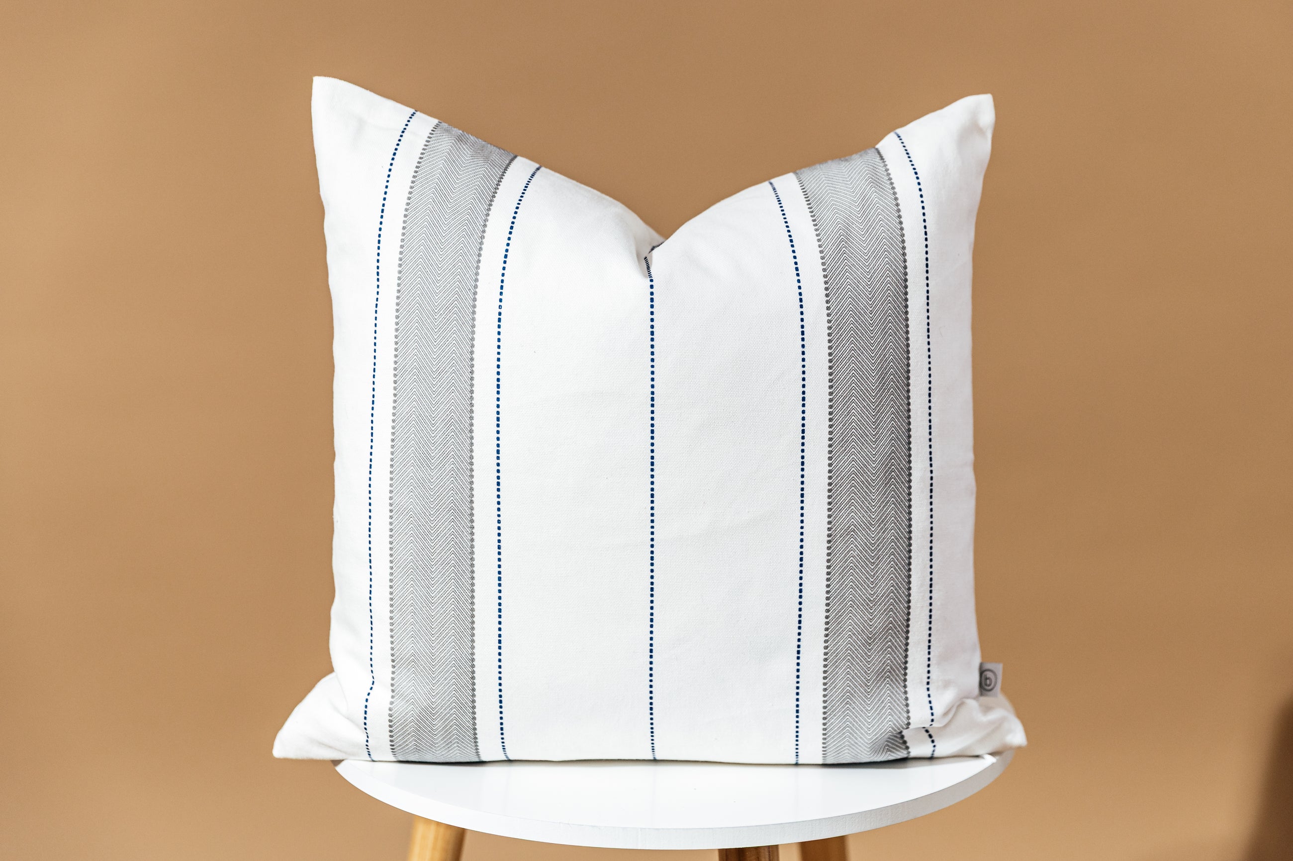 A white, gray, and navy striped pillow sitting on a stool in front of a brown backdrop.