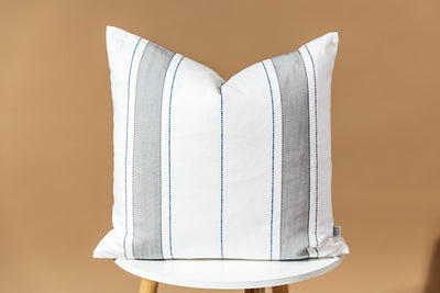 A white, gray, and navy striped pillow sitting on a stool in front of a brown backdrop.