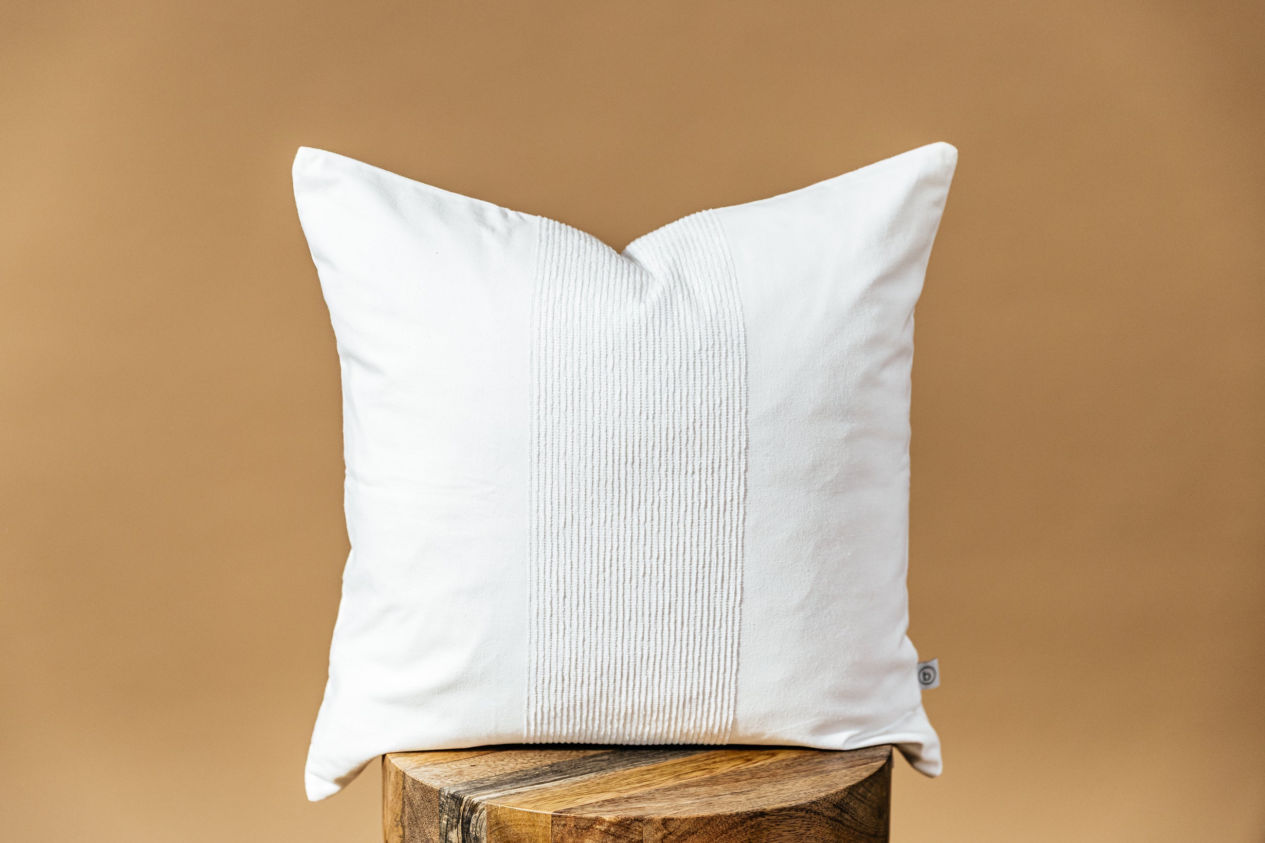 A white throw pillow on a wood stool over a brown backdrop.