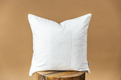 A white throw pillow on a wood stool over a brown backdrop.
