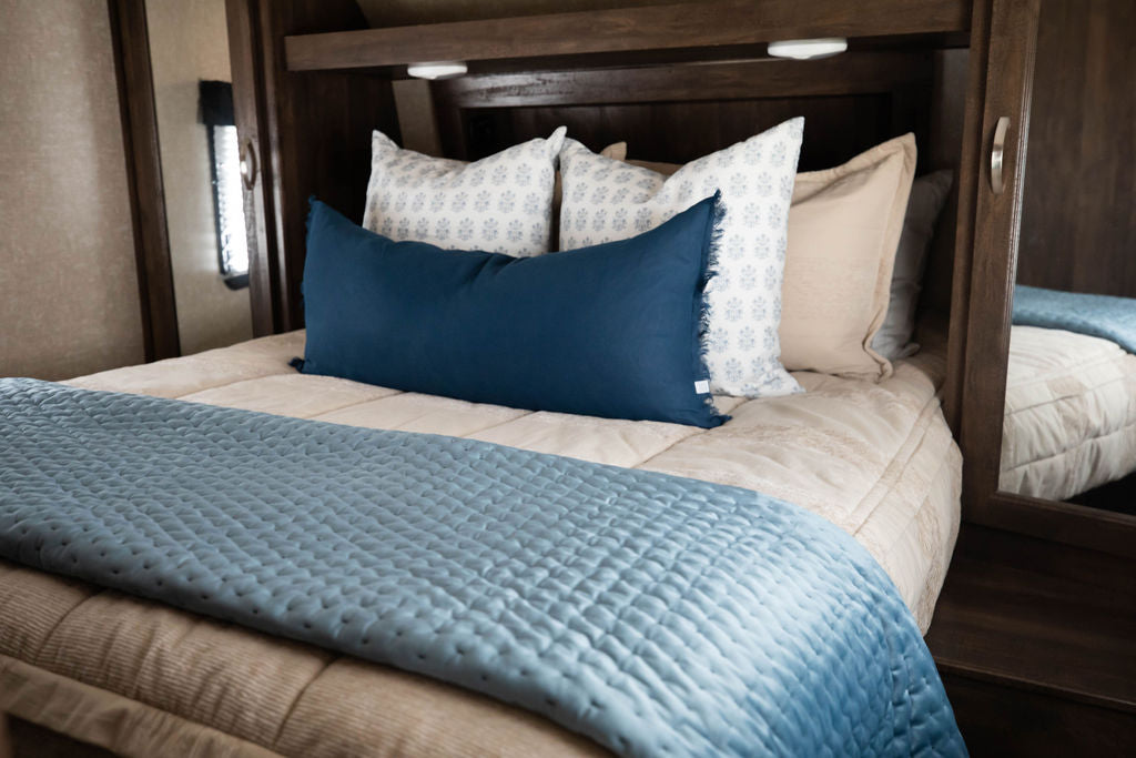 A bed in an RV made with tan faded stripes zipper bedding and an assortment of neutral and blue pattern throw pillows and blanket.