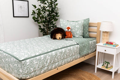 A twin bed made with sage green forest animal kids zipper bedding with a nightstand beside the bed holding books and a lamp.