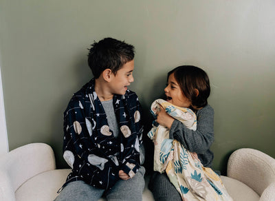 A brother and sister sitting beside each other and laughing together, both wrapped up in children's blankets.