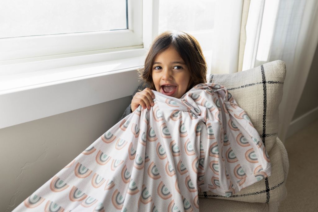 A little girl sitting on a sun bench underneath a rainbow printed child's blanket.