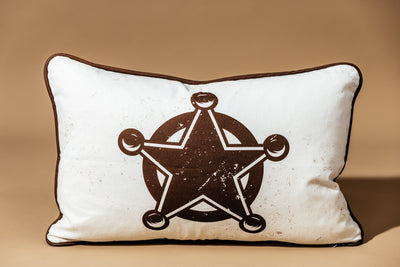 A western pillow with a sheriff style star in front of a brown backdrop.