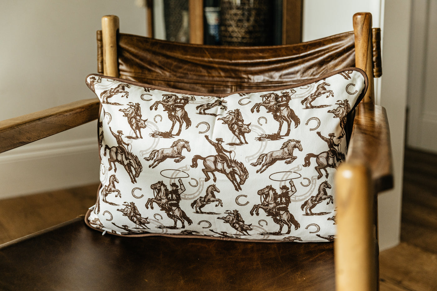 A western pillow with a print of cowboys, horses, and horseshoes sitting on a leather chair in a living room.