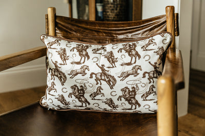 A western pillow with a print of cowboys, horses, and horseshoes sitting on a leather chair in a living room.