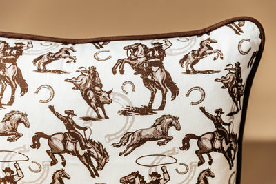 A close up shot of a western pillow with a printof cowboys, horses, and horseshoes, arranged in front of a brown backdrop.