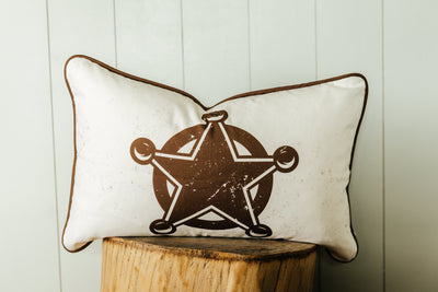 A western pillow with a sheriff style star on the front sitting on a wood stool in front of a white backdrop.