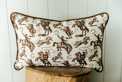 A western pillow with a print of cowboys, horses, and horseshoes, the pillow sitting on a wood stool in front of a white backdrop.