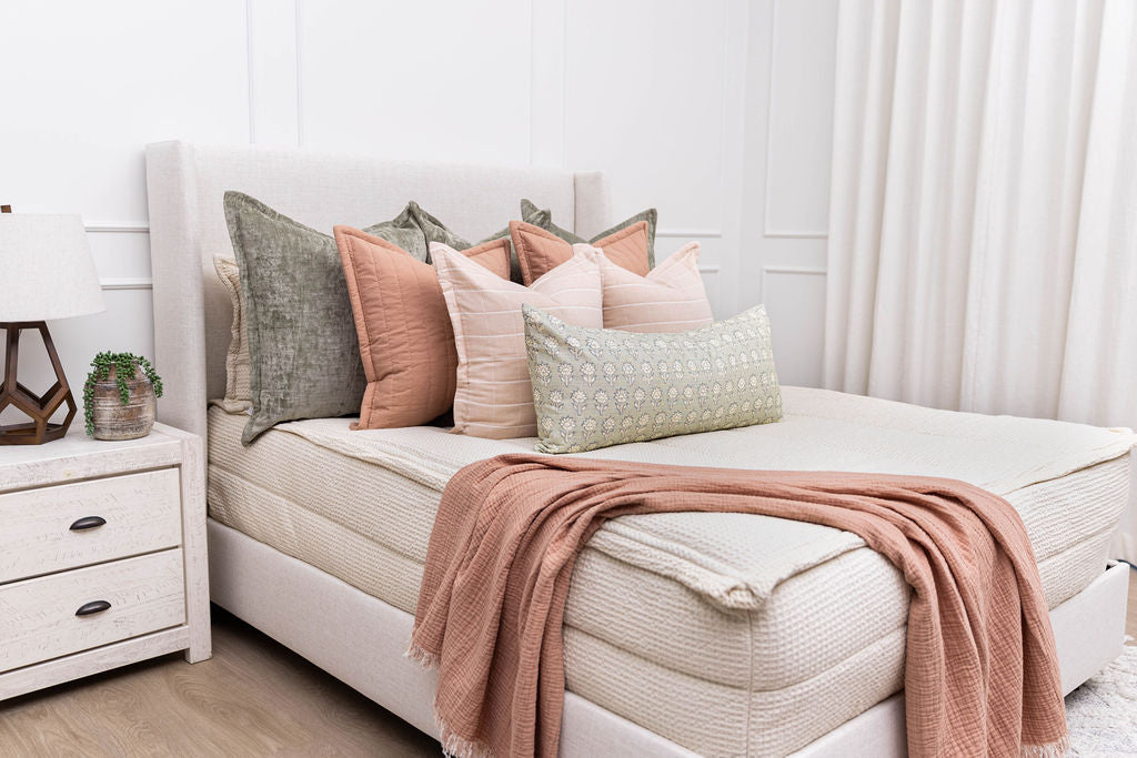 A brightly lit bedroom with a bed made with cream waffle textured zipper bedding an an assortment of green, terracotta pink, and floral pillows and blanket.