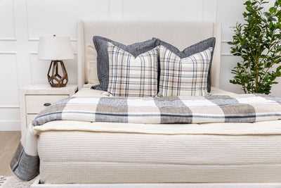 A bed made with cream waffle textured zipper bedding and an assortment of neutral plaid and dark gray pillows and blanket.