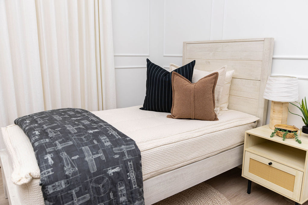 A twin bed made with cream waffle textured zipper bedding and an assortment of neutral black and brown pillows and an airplane printed blanket.