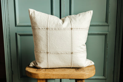 A plaid pillow on a wood stool in front of a green wall.
