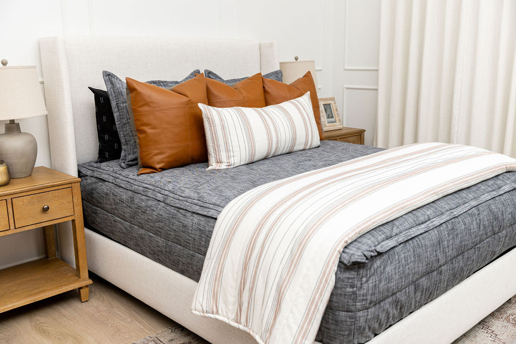 A queen bed made with dark charcoal gray zipper bedding and leather and neutral striped pillows and blanket.