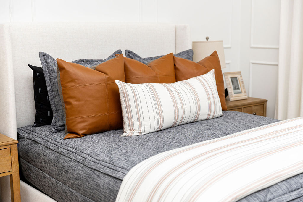 A close up view of a bed made with dark charcoal gray zipper bedding and leather and neutral striped pillows and blanket.