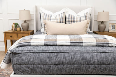 A bed made with dark charcoal gray zipper bedding and an assortment of neutral and plaid throw pillows and blanket.