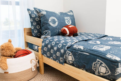 A toddler bed made with blue sports themed kids zipper bedding with a basket of stuffed animals beside it