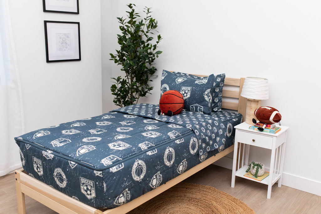 A twin bed made with blue sports themed kids zipper bedding and a nightstand beside it holding a lamp and a stuffed football