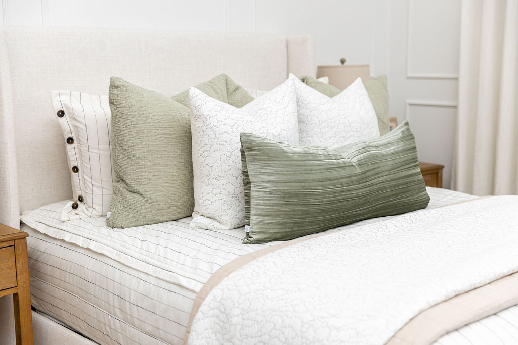 A brightly lit bedroom with a bed made with white and pinstripes zipper bedding and an assortment of neutral and gren pillows and blankets.