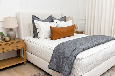 A brightly lit bedroom with a bed made with white and pinstripes zipper bedding and an assortment of neutral and dark charcoal gray pillows and blanket.