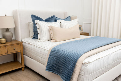 A brightly lit bedroom with a bed made with white and pinstripes zipper bedding and an assortment of neutral and velvet blue pillows and blankets.