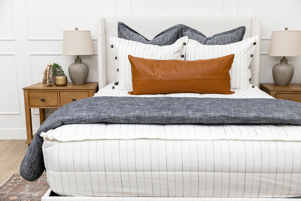 A bed made with white and pinstripes zipper bedding and an assortment of neutral and dark gray pillows and blanket.