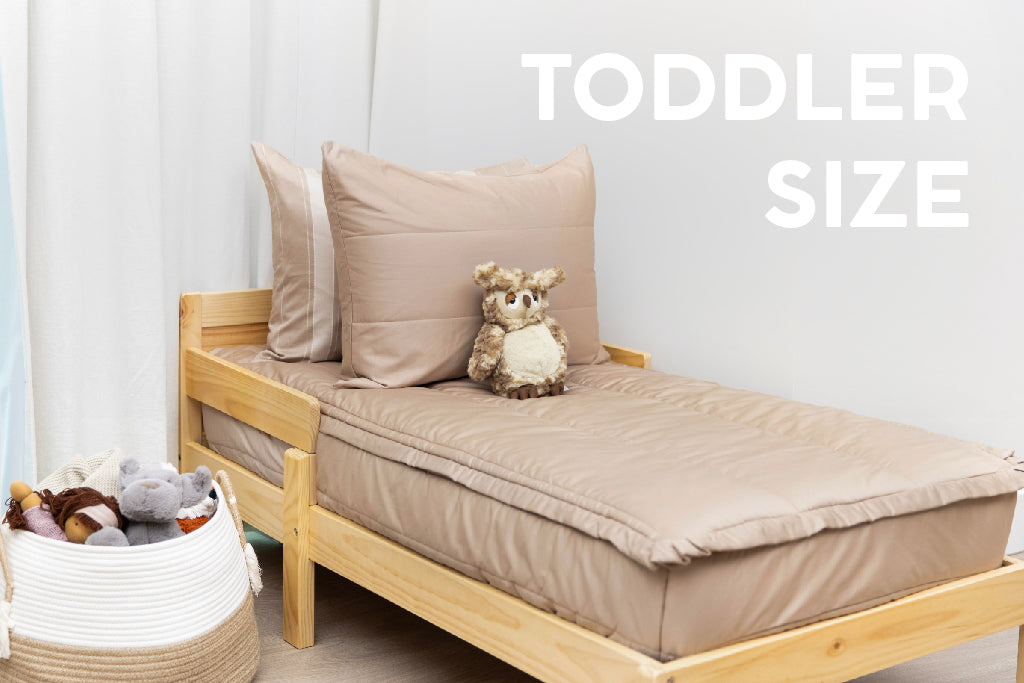 A toddler bed made with tan zipper bedding and a basket right next to the bed full of stuffed animals.