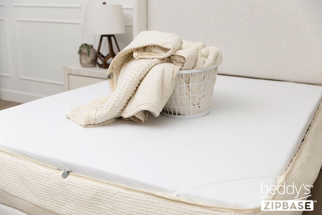 A bed made with just the fitted bedding base around the sides of the mattress, with the top comforter and ZipBase sheet in a basket on top of the bed.