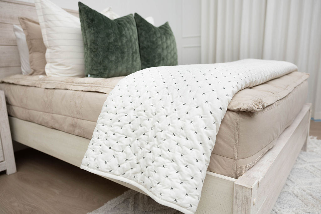 Creamy white blanket with green "x" stitching sitting on tan neutral zipper bedding