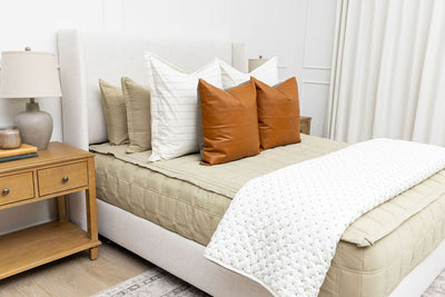 A queen bed in a brightly lit bedroom made with khaki green zipper bedding and an assortment of cream and leather bedding accessories.