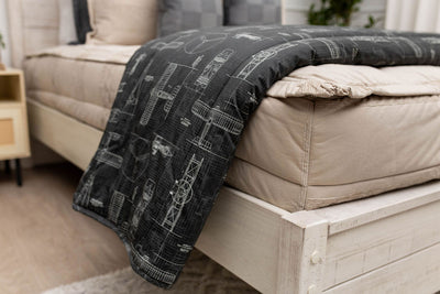 A side view of a bed made with tan zipper bedding with an airplane pattern black blanket folded over the end of the bed.