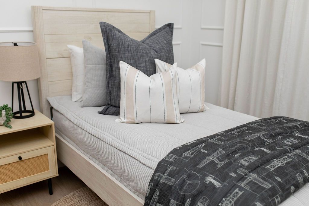 A twin bed made with light gray waffle textured zipper bedding and an assortment of neutral throw pillows and an airplane pattern black blanket.