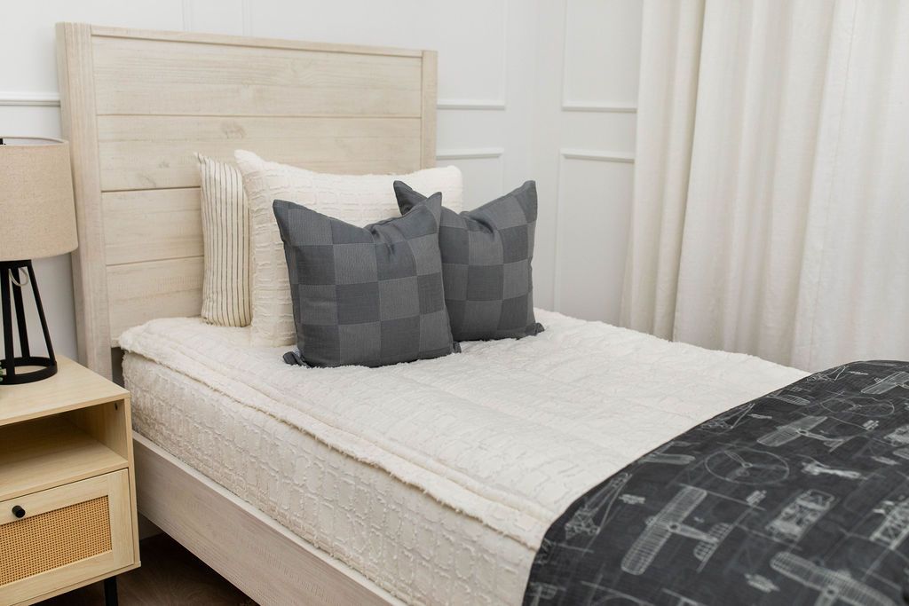 A twin bed made with cream textured zipper bedding and an assortment of neutral throw pillows and an airplane pattern black blanket.