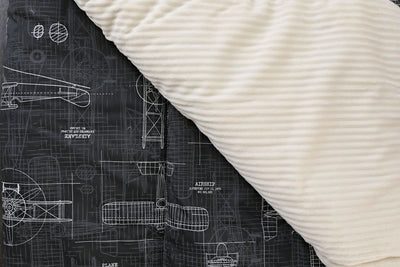 A charcoal airplane patterned blanket folded over to reveal the cream minky side.