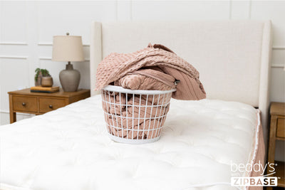 A bed made with just the fitted bedding base around the sides of the mattress, with the top comforter and ZipBase sheet in a basket on top of the bed.