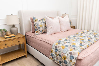 A brightly lit bedroom with a bed made with woven pink zipper bedding and an assortment of pink and floral tan pattern pillows and blanket.